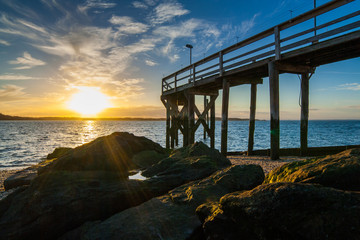 Obraz premium The setting sun with clouds in the background with a rocky beach, large algae-covered stones, and a wooden pier in the foreground. West Neck Beach in Huntington, NY, USA.