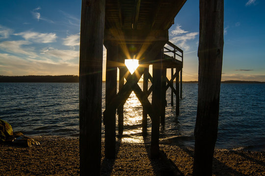 The setting sun shines between wooden planks underneath a pier at West Neck Beach in Huntington, NY, USA.