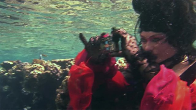 Young underwater model free diver in red dress photographs on camera in Red Sea. Filming a movie. Actress smiling at camera. Extreme sport in marine landscape, coral reefs, ocean inhabitants.