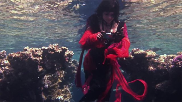 Young girl free diver in red dress photographs on camera underwater in Red Sea. Filming a movie. Extreme sport in marine landscape, coral reefs, ocean inhabitants.