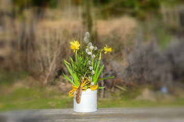 Ostergesteck, kleines Blechtöpfchen mit Bast und Feder, bepflanzt mit gelben Osterglocken, Heidelbeerästen und Palmkätzchen im Garten, weichgezeichnet
