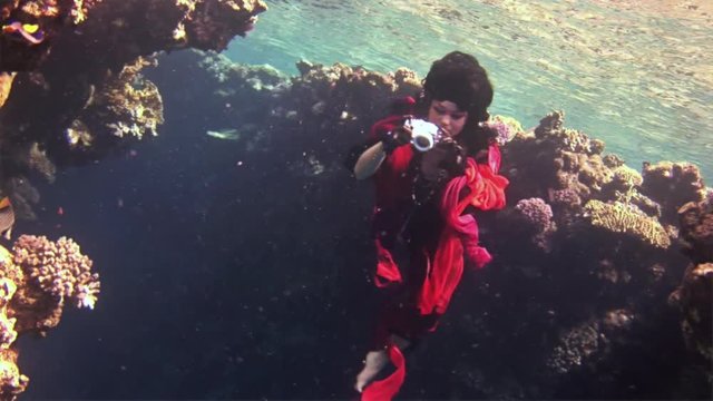 Young underwater model free diver in red dress photographs on camera in Red Sea. Filming a movie. Extreme sport in marine landscape, coral reefs, ocean inhabitants.