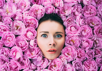 Portrait of beautiful young woman with pink lips and green eyes among the pink roses, wondering emotion