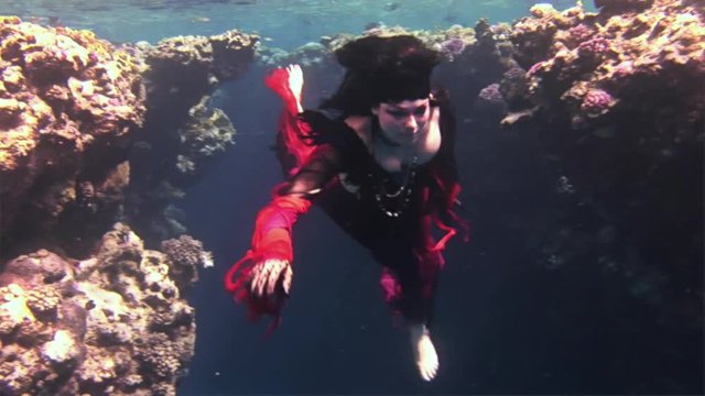 Underwater model free diver in red dress on background of corals in Red Sea. Filming a movie. Young girl smiling at camera. Extreme sport in marine landscape, coral reefs, ocean inhabitants.