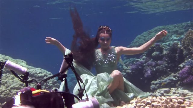 Underwater model free diver poses for camera on background of corals in Red Sea. Filming a movie. Young girl smiling. Extreme sport in marine landscape, coral reefs, ocean inhabitants.