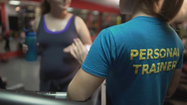 Fitness trainer consulting overweight lady exercising in gym, weightloss control