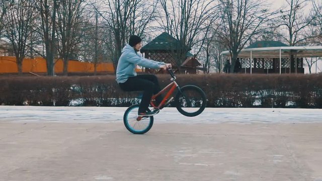 Teenager doing tricks, rides the BMX on the rear wheel in the winter park. Slow motion. 120 fps.