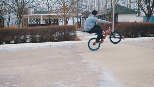 Teenager doing tricks, rides the BMX on the rear wheel in the winter park. Slow motion. 120 fps.