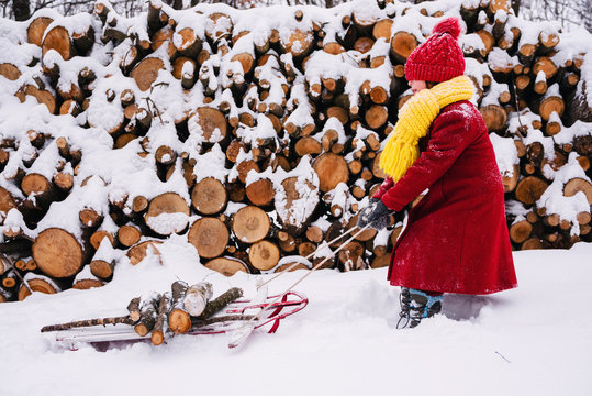 Side view of a young Girl in a winter coat, hat, scarf and gloves standing in the snow pulling a sledge laden with firewood
