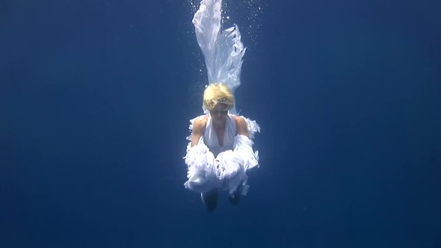 Young girl model underwater white angel costume on blue background in Red Sea. Filming a movie at camera. Extreme sport in marine landscape, coral reefs, ocean inhabitants. Free diving.