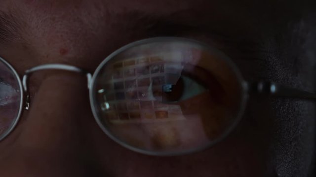 Surfing internet with screen reflection in glasses. Man in glasses is surfing web internet in a dark room. Closeup candid face shot.