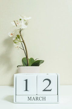 Closeup white wooden calendar with black 12 march word with white orchid flower on white wood desk and cream color wallpaper in room textured background , selective focus at the calendar