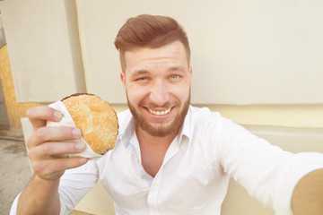 Hungry businessman making selfies with hamburger during his lunch time. Smiling short-haired man resting outdoors. Toned image.