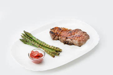 Grilled beef steak with asparagus and red sauce on a white plate, over white background