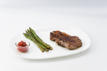 Grilled beef steak with asparagus and red sauce on a white plate, over white background
