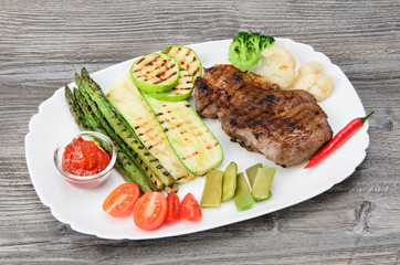 Grilled beef grilled with asparagus, zucchini, lobi, broccoli, tomato, chili and red sauce on a white plate, over wood background
