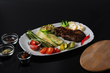 Grilled beef grilled steak with asparagus, zucchini, lobi, broccoli, tomato, chili and red sauce on a white plate, on a black glossy background