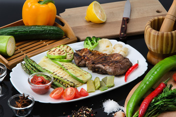 Grilled beef grilled steak with asparagus, zucchini, lobi, broccoli, tomato, chili and red sauce on a white plate, on a black glossy background
