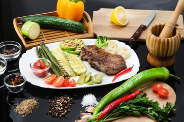 Grilled beef grilled steak with asparagus, zucchini, lobi, broccoli, tomato, chili and red sauce on a white plate, on a black glossy background