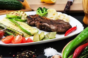 Grilled beef grilled steak with asparagus, zucchini, lobi, broccoli, tomato, chili and red sauce on a white plate, on a black glossy background
