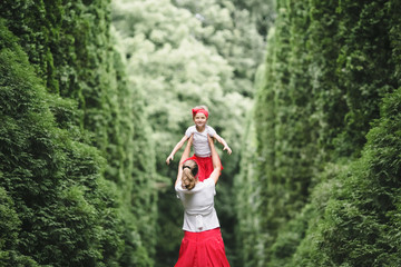 Woman holds her little daughter up standing with her among tall green bushes