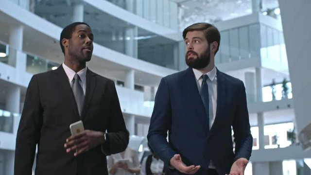 Dolly shot of African businessman in suit walking through lobby of modern business center and discussing work with gesturing bearded colleague 