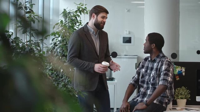 Tilt up of bearded businessman in casual outfit standing in office lounge area and chatting with male African colleague during lunch break 