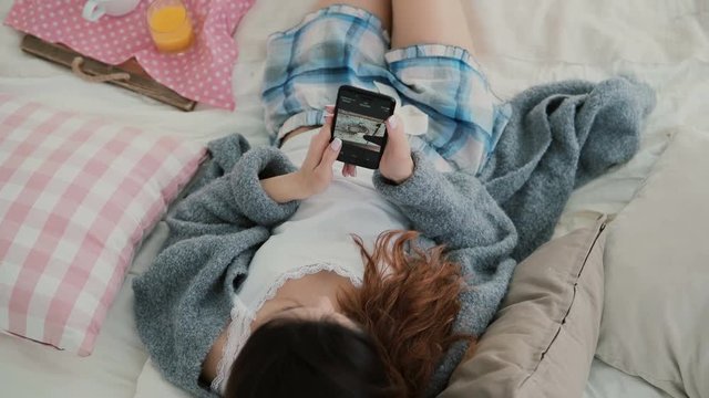 Young brunette woman lying on a bed. Beautiful girl using smartphone during breakfast. Female watches photos from trip.