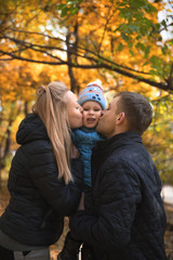 Fototapeta premium Young family sitting in park or forest. Father and mother kissing their small cute son