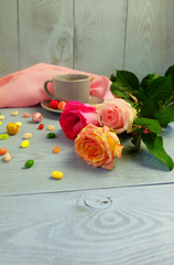 A bouquet of roses, a cup of tea, a pink napkin, colorful candy on a dyed wooden background.