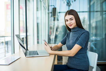 Side view of young smiling businesswoman sitting laptop at table in coffee shop and uses smartphone.