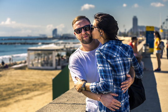 Portrait Of Young And Attractive Couple Close Up Standing Next To The Sandy Beach. Woman Is Kissing Her Man In Cheek. Barcelona, Spain