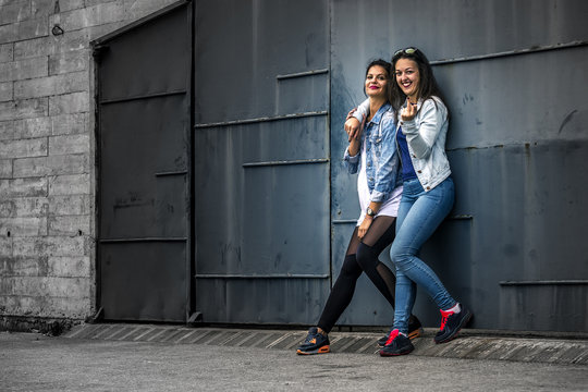 Portrait of two young and attractive women standing next to the wall, looking at camera. Barcelona, Spain. One girl is showing the middle finger at camera.