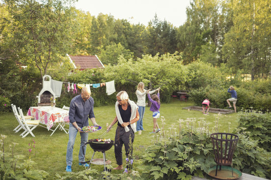 Man and woman cooking vegetables on barbecue grill with kids enjoying garden party