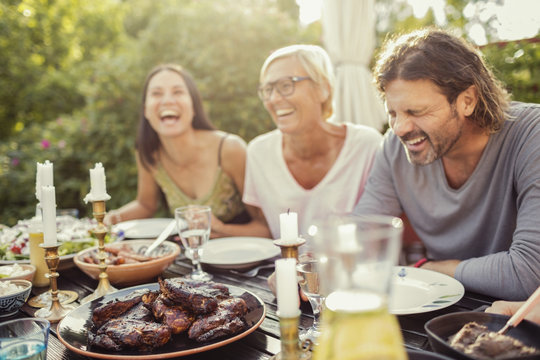 Cheerful couple and female friend laughing on dining table during garden party in back yard