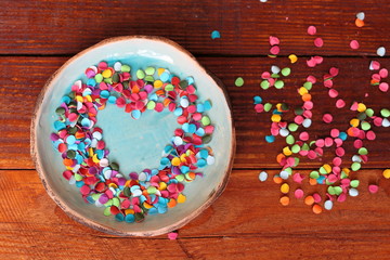 Heart from multicolored confetti on small turquoise handmade ceramic plate on background of wooden boards. Bright true love and romance