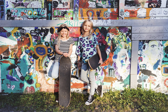 Girls holding skateboards, standing against graffiti wall