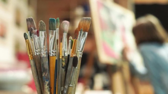 Set of paintbrushes in the foreground and girl painting a picture on the easel in art studio on the background