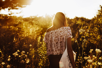 stylish hipster woman with eco bag walking in sunlight among wildflowers in summer evening field under sun rays. atmospheric moment. boho girl relaxing in meadow. vacation and travel
