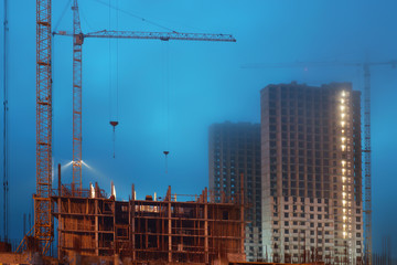 Fototapeta na wymiar Cranes on a large construction site, unfinished houses, fog covers the upper floors, evening twilight