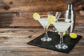 Alcoholic cocktail - gin tonic, martini, margarita. Decorated with slice of lime, with a shaker on a black slate on a wooden table, copy space