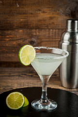 Alcoholic cocktail - gin tonic, martini, margarita. Decorated with slice of lime, with a shaker on a black slate on a wooden table, copy space, vertical