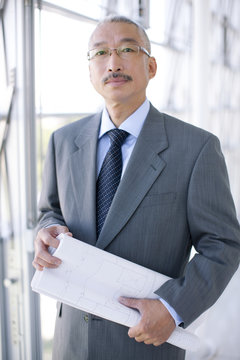 Portrait of businessman standing by window, holding blueprint