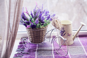 Small decorative bicycle with wicker basket pour of spring lavender bouquet and white watering can on the windowsill covered lilac plaid. Close-up - obrazy, fototapety, plakaty