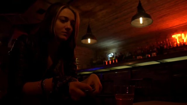 Beautiful nice girl in restaurant is holding a mobile phone. Bar stand with drinks, menu, sign, glass. Red light and glow. Slow motion
