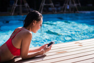 Beautiful woman leaning on poolside and typing a text message on cellphone