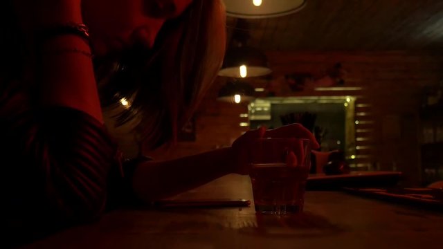 Pretty nice girl in restaurant holding a glass and then corrects hair. Bar stand with drinks, menu, sign, glass. Red light and glow. Sad girl waiting near the bar. Slow motion