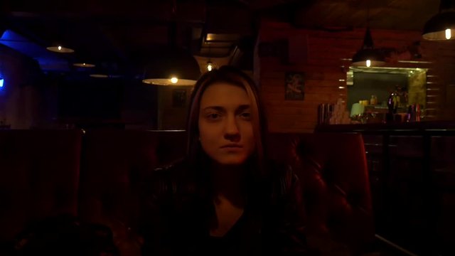 Sad beautiful nice girl in a restaurant. Bar stand with drinks, menu, sign, glass. Red and blue light and glow. Leather sofa. Slow motion