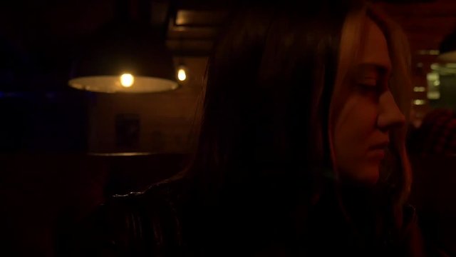 Sad nice girl in a restaurant. Bar stand with drinks, menu, sign, glass. Red and blue light and glow. Leather sofa. Turning to camera. Close shot. Slow motion
