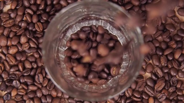 Coffee beans are poured into a glass beaker close up. Grains are poured over the edge. Slow motion.